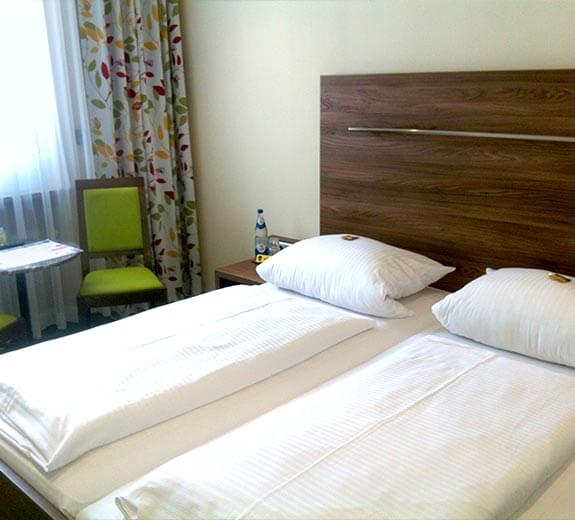 Helles Zimmer mit zwei Betten, einem Nachttisch und einem kleinen Tisch mit Stühlen an einem Fenster.