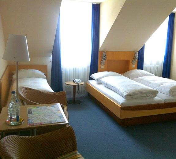 Ein helles Zimmer mit zwei Betten, einem Tisch, einem Stuhl und einem Fenster mit Vorhängen.